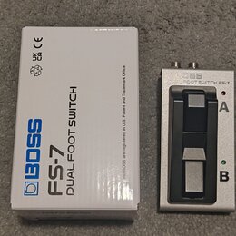 Bílá krabice s modrým nápisem BOSS FS-7 DUAL FOOT SWITCH a doprovodnými certifikačními symboly. Vedle krabice je stříbrný dvoupedálový spínací jednot…