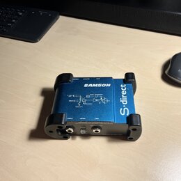 Modrá DI box s černými gumovými nárazníky na rozích. Na horní straně je bílý nápis SAMSON a schéma zapojení s texty INPUT, PAD, HIGH Z, BATTERY PHANT…
