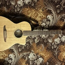 Akustická kytara s vyřezem v těle, přírodní dřevěnou barvou a tmavým hmatníkem. Na hlavě je viditelné logo Fender. Kytara má černý popruh a hnědý kob…