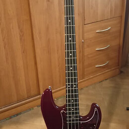 Tmavě červená elektrická basová kytara s tmavým hmatníkem a červeně vzorovaným pickguardem. Na hlavě nástroje je viditelné logo "Music Man". Kytara s…