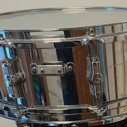 Kovový snare drum s chromovaným, vysoce lesklým povrchem. Na kotečích jsou viditelné loga ve tvaru stylizovaného písmena „S“. Nástroj vykazuje drobné…