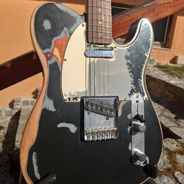 Elektrická kytara typu Telecaster s černým lakem, který je výrazně opotřebovaný a v mnoha místech odloupaný, čímž odhaluje světlé dřevo. Nástroj má k…