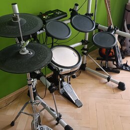 Elektronická bicí souprava s černými gumovými pady a činely na stříbrném kovovém rámu. Na bílém bubnu je viditelný nápis OXO. Na pedálu basového bubn…