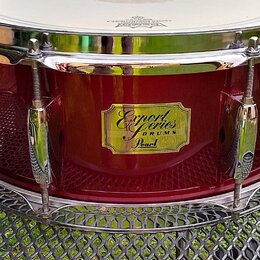 Tmavě červený buben s kovovými obručmi a napínacími šrouby. Uprostřed korpusu je zlatý nápis Export Series DRUMS Pearl. Povrch nástroje je lesklý a v…