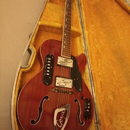 Polodupatá elektrická kytara v červeném provedení s f-otvory, položená v otevřeném kufru s žlutým vnitřkem. Na těle je černý pickguard a kovový ozdob…
