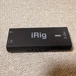 Černý obdélníkový audio rozhraní s nápisem iRig a označením HD 2. Na levé straně je vidět symbol mikrofonu a ikona baterie. Povrch zařízení je matný,…