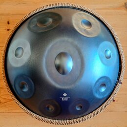 Kulatý kovový handpan s modravým, gradientním povrchem a několika vyhlubecinami (tóny). Nástroj má pletený lem v béžové barvě. V dolní části je bílý…