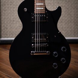 Černá elektrická kytara s tvarem single-cutaway a dvěma humbuckery. Na hrazdiku v pozadí je vidět bílý nápis Marshall. Kytara má čtyři ovládací knofl…