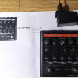 Černý efektový pedál s oranžovým pruhem, na kterém je nápis TC-HELICON harmonyM a VOICETONE™. Zařízení obsahuje několik otočných potenciometrů, tlačí…