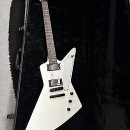 Bílá elektrická kytara s ostrou hranatou formou těla, položená v černém tvrdém kufru. Na hmatníku jsou dva humbuckery a tři ovládací knoflíky, tělo j…