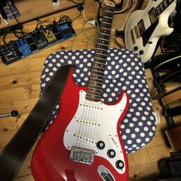Červená elektrická kytara s bílým pickguardem a hnědým hnedým popruhem. Na hlavě kytary je viditelné logo Squier. Kytara má tři snímače, pětiknoblové…