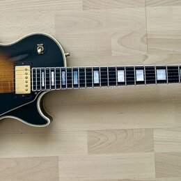 Elektrická kytara s tónem sunburst a černým povrchem na okrajích. Na hlavě je logo Gibson a nápis Les Paul Custom. Nástroj má zlatý hardware, dva hum…