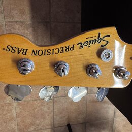 Fotografie zachycuje hlavu basové kytary z přírodního dřeva s čtyřmi chromovanými ladičkami. Na povrchu je černý nápis Squier BY FENDER a PRECISION B…