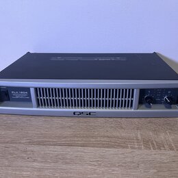 Stříbrný rackový výkonový zesilovač s černým horním krytem. Na předním panelu je nápis PLX 1804 PROFESSIONAL POWER AMPLIFIER a v dolní části logo QSC…