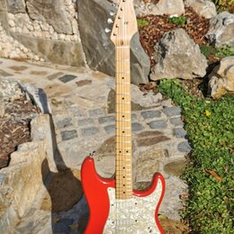 Červená elektrická kytara typu Stratocaster s bílým pickguardem a světlou hnedou hmatníkovou deskou. Na hlavě kytary je viditelné logo Fender. Nástro…
