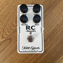 Bílý kytarový efekt s nápisy RC -booster a Xotic Effects CALIFORNIA. Na horní straně jsou čtyři černé otočné knoflíky s popisky GAIN, TREBLE, VOLUME…