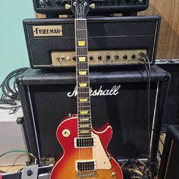 Elektrická kytara s tvarem singlecut a povrchovou úpravou sunburst v červeně a žluté. Na hlavě je bílé logo Gibson a nápis Les Paul Custom. Kytara má…