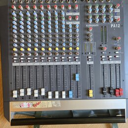 Šedý mixážní pult s nápisem ALLEN&HEATH a označením modelu PA12. Zařízení obsahuje řadu vertikálních faderů, barevné otočné knoflíky a různé typy vst…