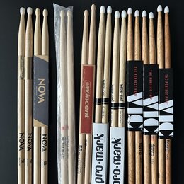 Sada různých bubnových paliček z přírodního dřeva rozložená na černém povrchu. Jsou zde paličky s nápisy NOVA, Vincent, Pro-Mark a Vic Firth (označen…