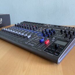Šedý mixážní pult s modrými prvky a řadou posuvníků. Na pravé straně je nápis L-12 MultiTrak a logo ZOOM, vedle nich se nachází displej. Zařízení je…