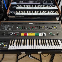 Hnědý syntetizátor značky YAMAHA s klaviatura a ovládacím panelem s různobarevnými tlačítky. Nástroj je umístěn v černém přepravním kufru a stojí na…
