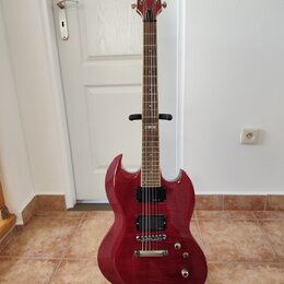 Červená elektrická kytara s dvojstupňovitým tělem a hnědým hmatníkem, umístěná na černém stojanu. Na hlavě nástroje je bílé logo Ltd. Kytara má dva h…