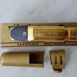 Zlatý mosazný saxofonový mouthpiece s černým náústkem, položený na kartónové krabičce. Na mouthpiece je vyryt nápis "Otto Link". Krabička obsahuje te…