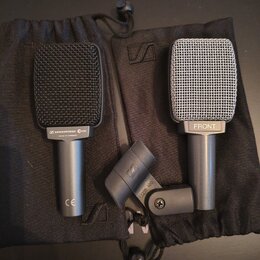 Dvě mikrofony s mřížkou a jeden držák v černých látkových pytlích. Levý mikrofon je černý s nápisem SENNHEISER e606 a MADE IN GERMANY, v dolní části…
