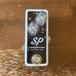 Kompresorový pedál s bílým rámem a černým horním panelem. Na panelu je bílý nápis „SP compressor BY xotic usa“. Pedál má dva průhledné ovládací knofl…