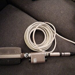 Stříbrný mikrofon s kovovou mřížkou a připojeným bílým kabelem. Na těle mikrofonu je viditelný nápis: „AKG“, „MADE IN AUSTRIA“, „VIENNA“. Povrch vyka…