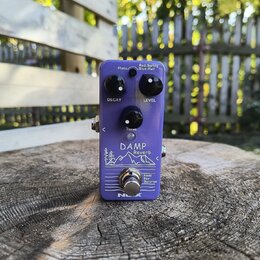 Fialový kytarový efekt s nápisem DAMP Reverb a grafickým motivem hor. Na horní straně jsou tři černé otočné oválné knoflíky s popisky DECAY, LEVEL a…