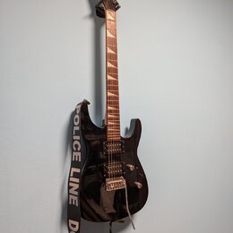 Černá elektrická kytara s hnědým hmatníkem a dvěma humbuckery. Na hlavě nástroje je viditelné logo Schecter. Kytara má nasazený černobílý popruh s ná…
