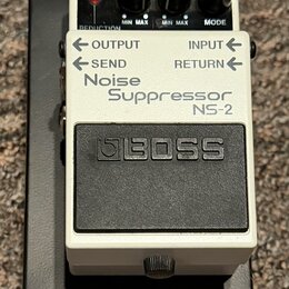 Bílý efekt pedál s černým spínacím tlačítkem a nápisem BOSS. Na přední straně je text Noise Suppressor NS-2 a označení vstupů OUTPUT, INPUT, SEND a R…