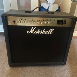 Černý kytarový kombo zesilovač s látkovou mřížkou. Na přední straně je bílé logo Marshall a na ovládacím panelu nápis MG 100W. Panel obsahuje řadu ot…