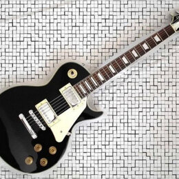 Černá elektrická kytara s bílým pickguardem a dvěma humbuckery. Na hlavě je viditelné logo Gibson. Nástroj vykazuje známky běžného používání, povrch…