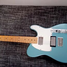 Modrá elektrická kytara s bílým pickguardem a světlou hnedou hmatníkovou deskou. Na hlavě je viditelné logo Fender. Kytara má dva snímače a kovový mo…
