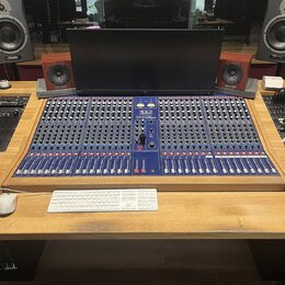 Na dřevěném stole je umístěno velké modré míchací pult s nápisem TLA Audio M800. Nad pultem stojí široký počítačový monitor a dva dřevěné studiové mo…