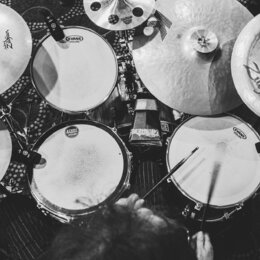 Černobílá fotografie z pohledu bubeníka na bicí sadu. Na čínalech jsou viditelná loga Zildjian a Sabian, na blanách bubnů nápisy EVANS a ETAMA. Sníme…