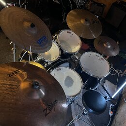 Celá bicí souprava s černými kotly a kovovými obručmi. Na činelech jsou viditelné nápisy Zildjian, ZBT a VFIP. Na bubnech je vidět logo TAMA. Souprav…