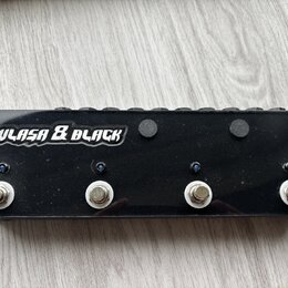 Černý obdélníkový pedalboard s nápisem ULASA & BLACK v bílém stylizovaném písmu. Na přední straně jsou čtyři kovové spínací tlačítka s bílými podložk…