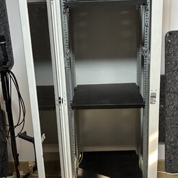 Bílá kovová serverová skříň s otevřenými dveřmi. V interiéru jsou umístěny tři černé police na různých úrovních. Povrch skříně vykazuje běžné známky…