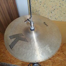 Kruhové činely z mosazného kovu s viditelným broušením povrchu. Na ploše je černý grafický symbol šipky a nápisy „Hi Hat TOP“ oraz „14" 105Hz L21“. V…