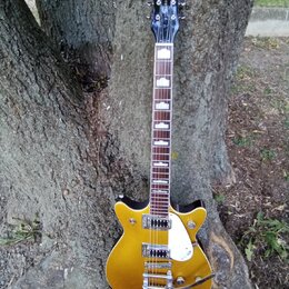 Zlatá elektrická kytara s černým hmatníkem a bílými vložkami. Na hlavě je nápis GRETSCH, na těle se nachází bílý plechtrguard a nápis GRETSCH. Nástro…