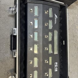 Černý MIDI kontrolér s dvanácti stisknutelnými spínači, z nichž každý má vedle malý displej. Na pravé straně je nápis FRACTAL AUDIO SYSTEMS a FC-12 F…