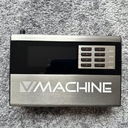 Kovové zařízení stříbrné barvy s černým displejem. Na spodní části je bílý nápis MACHINE s logem ve tvaru trojúhelníku. V pravé části jsou čtyři řady…