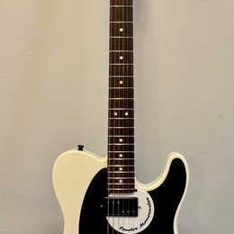 Bílá elektrická kytara s černým pickguardem a hnědým hmatníkem. Na hlavě je viditelné logo Fender a na pickguardu nápis Fender Mono-caster. Nástroj j…