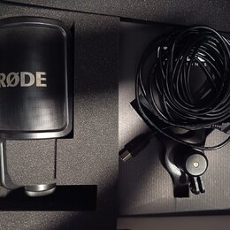 Černý mikrofon s nápisem RØDE umístěný v originální pěnové výplni. Vedle něj je srolovaný černý kabel USB a držák mikrofonu. Všechny komponenty vypad…
