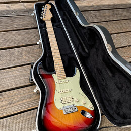 Elektrická kytara v barvě sunburst s bílým pickguardem, umístěná v černém tvrdém kufru. Na hlavě kytary je viditelné logo Fender. Nástroj má tři sním…