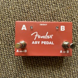 Červený kovový kytarový pedál s nápisem Fender ABY PEDAL. Na horní straně jsou bílá písmena A a B, nápis IN/OUT a dva kovové spínače s popisky A and…