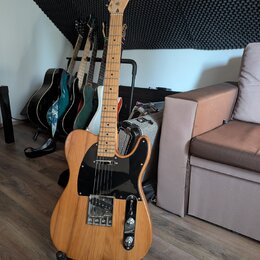 Elektrická kytara s přírodním dřevěným povrchem, černým pickguardem a chromovanými komponenty. Na hlavě je viditelné logo "Telecaster". V pozadí stoj…